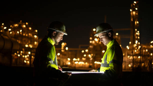 istock Engineer survey team wear uniform and helmet stand workplace checking blueprint project and radio communication inspection work construction site with night lights oil refinery background 2047190587
