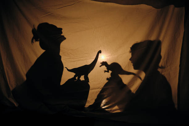 Silhouette of laughing mother playing with cute son and storytelling with brachiosaurus in shadow theater sitting behind sheet