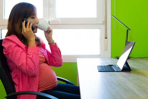 pregnant latina woman talks on her cell phone. In the office in front of her laptop.