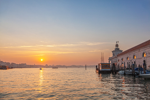 As the sun rises over the Venetian Lagoon in Venice, Italy, hues of golden and pink paint the sky, casting a warm glow over the tranquil waters.