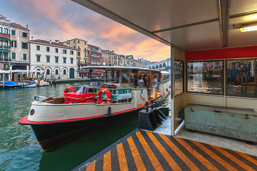 A vaporetto glides gracefully along the Grand Canal in Venice, Italy, its image blurred with motion as it arrives at the stop. In the background, the iconic waterway of Venice stretches majestically, flanked by historic palaces and buildings.