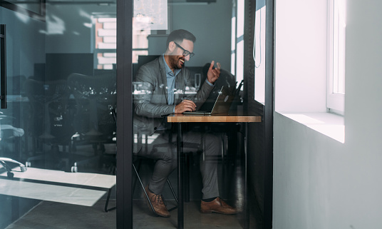 Shot of a businessman working in private cubicle at a coworking office. Confident businessman using silent pod room in modern co-working office. Businessman having important online conversation with customer. Shot of a man using a laptop for video conference. Smiling businessman in a video call using laptop in the office. The view is through glass.