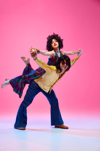istock Disco dancing couple in dynamic lift pose vibrant retro style outfit against gradient pink studio background. Energetic moves. 2047443745