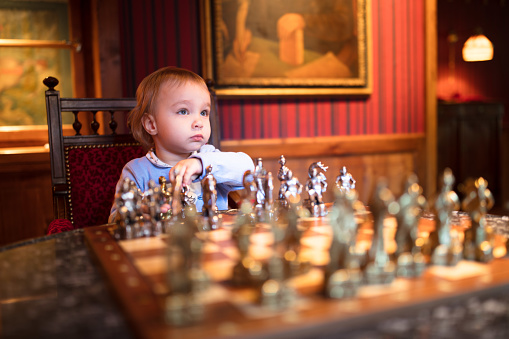 A cute little girl plays chess, carefully studying her next move in front of a beautiful chessboard. She sits in an elegant room draped in red.