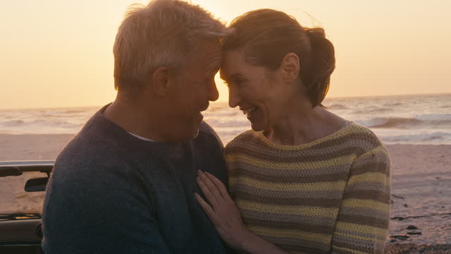 Portrait Of Retired Senior Couple On Vacation By Classic Sports Car At Beach Watching Sunrise