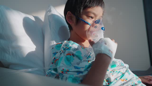 Asian boy on hospital bed with sunlight, holding an inhaler mask and looking outside the window, absent-minded
