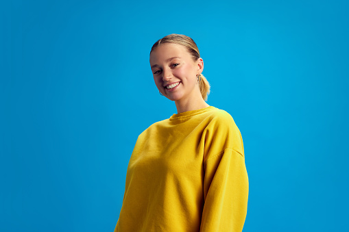 Portrait of beautiful young girl with ponytail, nude makeup, wearing yellow sweatshirt, smiling against blue studio background. Concept of youth, human emotions, casual fashion