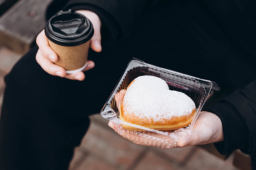 A paper cup of coffee and a heart-shaped donut in male hands, close-up. A man in a black jacket with a donut outside.