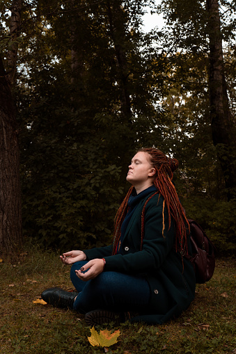 vertical portrait of adult woman with dreadlocks in casual wear sitting in forest and doing meditation