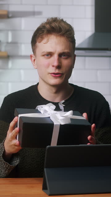 Vertical video. Young blonde vlogger man sitting in kitchen with tablet computer, catch the gift box in the air, advertising product , talking to camera, recording video