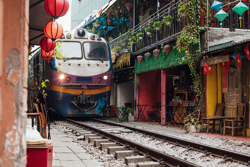 Train travelling through Hanoi, in the famous tourist location. It shows the extreme proximity to the shops and buildings on either side. Traditional colourful lanterns and flags decorate the buildings and cafes en route.