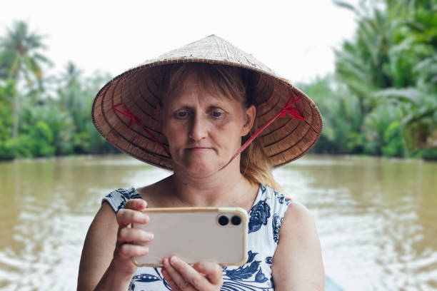 A  frontal close up view of a European single tourist, sat in a small canoe , taking photographs on her  smart phone, whilst an out of focus individual paddles her along in the rear of the boat. Either side of the river there is lush green vegetation and palm trees, the tourist is wearing a traditional triangular hat made od palm leaves, with a red chin strap.