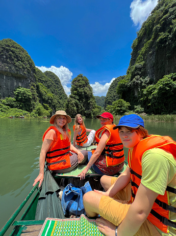 A group of Life jacket-wearing tourists are on a tourist row boat in Trang An, eco tourist site Vietnam. This family has with them,  teenagers and parents. This real family turn and smile at the camera, in the background, this film set location, is evidently beautiful. The karst limestone stacks have lush greenery down to the water. this family have engineered a customised vacation, away from the crowds.