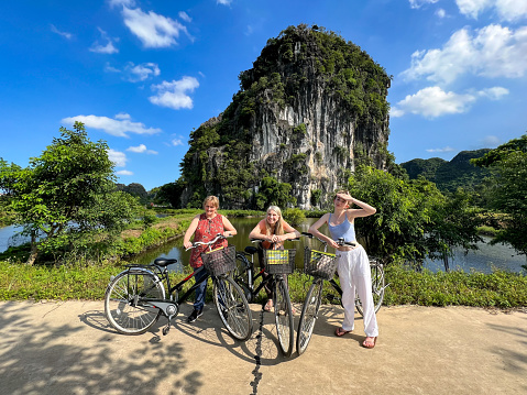 Three female family members, pose on bicycles infront of a limestone karst rock formation. These European tourists, are smiling and enjoying the waterways and lush tropical surroundings.