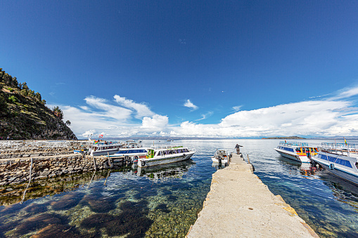 Sun Island, Bolivia - January 6, 2020: The harbor of Sun Island offers a picturesque scene, characterized by a stone walkway extending into the clear waters of Lake Titicaca. Cruise ships frequently dock nearby, adding to the vibrant atmosphere of the area. Against the backdrop of a clear blue sky, the tranquil and pristine waters of the lake provide a stunning contrast, enhancing the beauty of the surroundings. This serene setting invites visitors to take leisurely strolls along the waterfront, soak in the breathtaking views, and appreciate the natural splendor of Sun Island's harbor.