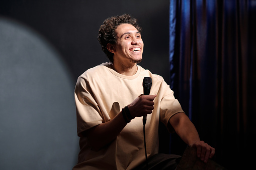 Young cheerful male comedian with microphone in hand performing new monologue in front of audience while sitting on stage of concert hall