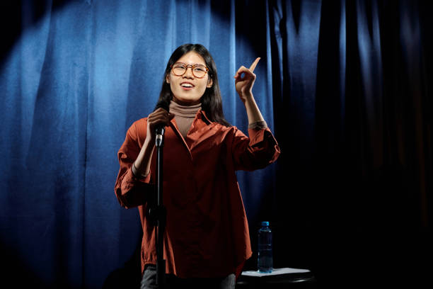 Young Asian woman with microphone Young Asian woman in eyeglasses and casualwear standing on stage with blue velvet curtains and speaking in microphone comedian stock pictures, royalty-free photos & images