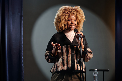 Young African American stand up comedian in wig speaking in microphone during performance while standing on stage in front of audience