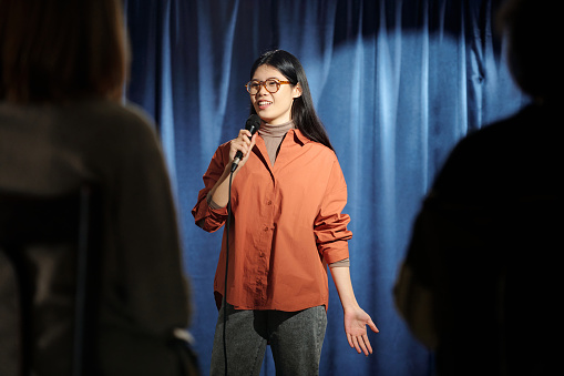 Young female comedian of stand up club standing on stage with blue curtains and pronouncing monologue in front of audience