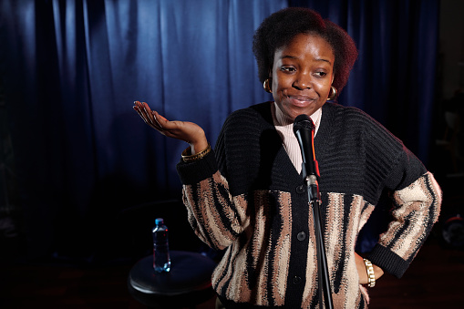 Young African American artist or actress of stand up club speaking in microphone while standing on stage with blue curtains