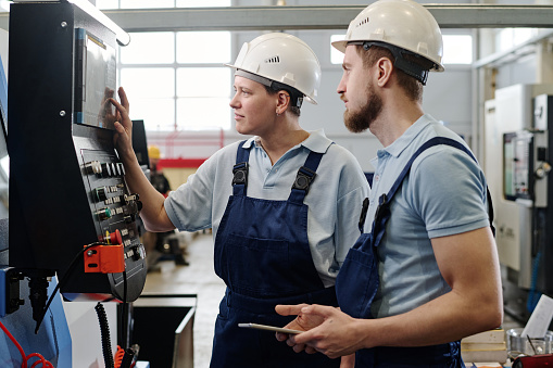 Side view medium shot of male and female engineers wearing hardhats working in factory using CNC machine