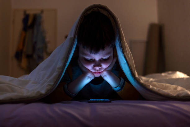 istock Boy playing games on mobile phone while lying on bed in bedroom under the blanket. He is hiding there. 2047776339