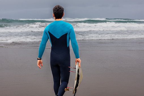 Back view of a male surfer in a blue wetsuit carrying a surfboard, heading towards the sea under a cloudy sky.