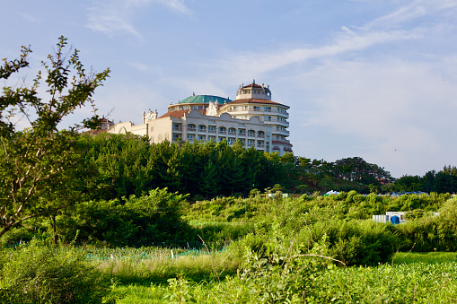 Yangyang County, South Korea - July 30th, 2019: Sol Beach Yangyang Resort is captured from afar, set against vibrant green fields under a blue sky, moments before sunset, highlighting its serene and picturesque setting.