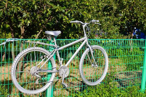 Yangyang County, South Korea - July 30th, 2019: An old, non-functioning bicycle, transformed into a piece of art by being painted entirely gray, hangs off a fence leading to fruit trees near Dongho Beach, symbolizing a creative repurpose.