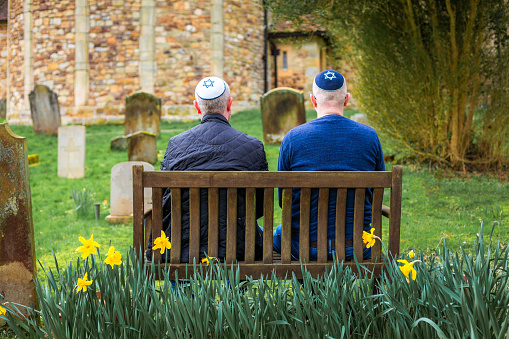Two Jewish men wearing skull caps (yarmulke) while sitting on a bench outside a synagogue. There are gravestones in the background. The men's skull caps have the Jewish symbol of the Star of David embroidered on them.