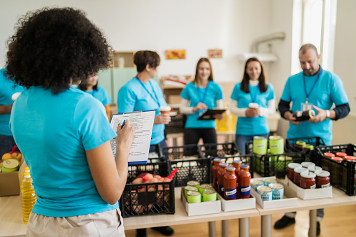 A woman overlooks a table full of donated groceries, checking her list while more charity workers take a coffee break in the background