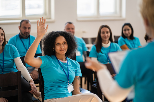 An African-Caribbean woman has her hand in the air to volunteer whilst sitting with her colleagues in a charity seminar
