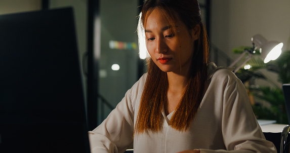 Closeup of focused young adult woman in a wheelchair working intently on a computer in an accessible office environment at night. ESG sustainable business office concept.