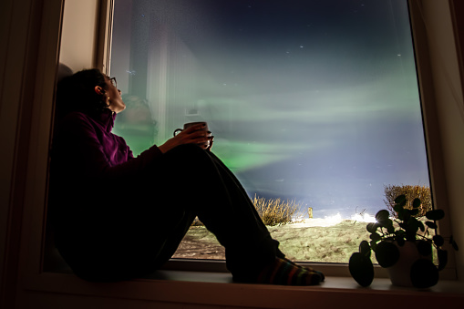Pretty young lady is looking  outside from window for northern lights at home.