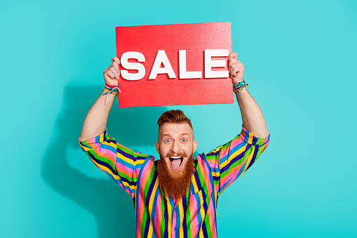 Portrait of handsome red hair bearded man in striped shirt hold red poster sale offer message isolated on aquamarine color background.