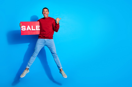 Full length portrait of handsome guy jump hold sale placard indicate finger empty space isolated on blue color background.