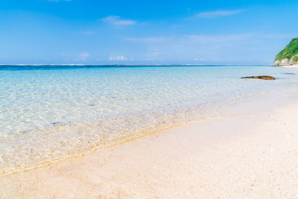 A serene tropical beach with crystal-clear blue waters gently lapping against the sandy shore. The lush green hillside, adorned with rocky outcrops, frames the scene. Bali - Melasti beach.