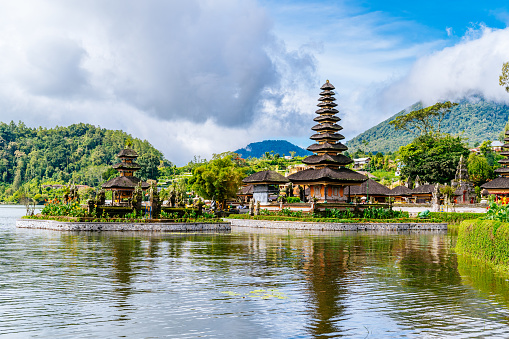 Pura Ulun Danu Bratan temple stands majestically with its multi-tiered shrine, surrounded by the reflective waters of Lake Bratan and lush greenery. The sky is partly cloudy, casting soft light on the serene landscape.