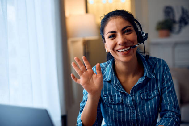 istock Happy Latin entrepreneur waving while working at her home office and looking at camera. 2047873336