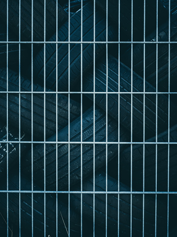 A lone, weathered tires rests behind a metal grid fence, illuminated by the faint glow of dim evening light which casts deep shadows around it, creating a stark contrast.