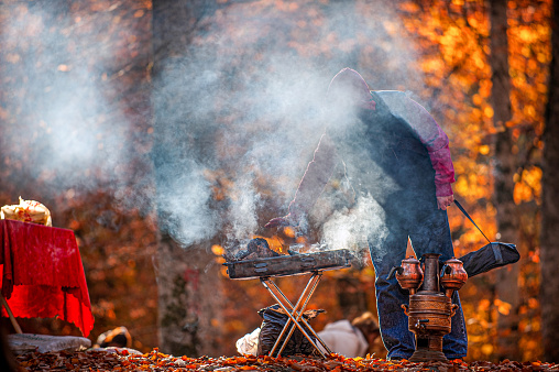 Man warming up over tea samovar and barbecue fire in the forest