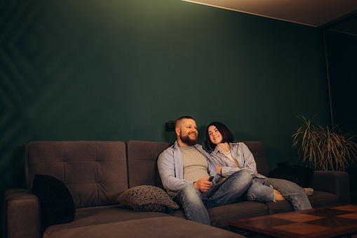 Couple Watches TV together while Sitting on a Couch in the Living Room. Girlfriend and Boyfriend embrace, cuddle, talk, smile and watch Television Streaming Services. Home with Cozy Stylish Interior.