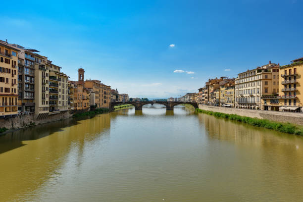 A captivating view of the Ponte Vecchio bridge with its characteristic shops lining the structure. The buildings along the Arno River add a vibrant contrast to the historical bridge, with their colorful facades and distinct architectural styles. The soft light and dynamic sky create a serene yet lively atmosphere in this iconic Florentine scene.