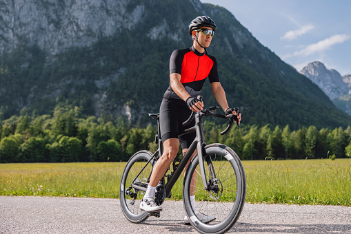 Professional male athlete about to go on a training bike ride to prepare for a race. His feet are on the ground and he is looking around. The man is wearing professional cycling gear including a helmet. In the background, there is a beautiful view of the mountainous countryside.