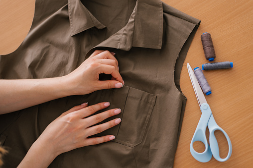 Close up view from above on the hands of a dressmaker, a young woman is manually working on the design of a new collection of khaki shirts. There are threads and scissors on the table nearby