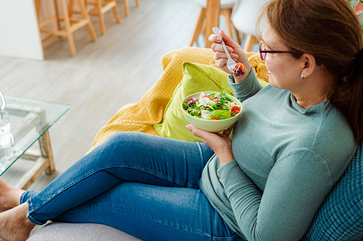 Mature woman eating salad relaxed on sofa. High resolution 42Mp indoors digital capture taken with SONY A7rII and Zeiss Batis 40mm F2.0 CF lens
