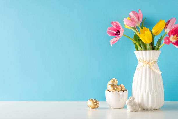 istock Spring sophistication theme. Side view shot of a table featuring themed bowl holding gold eggs, a porcelain rabbit, and a jar of vibrant tulips against a soft blue backdrop, leaving space for text 2047995739