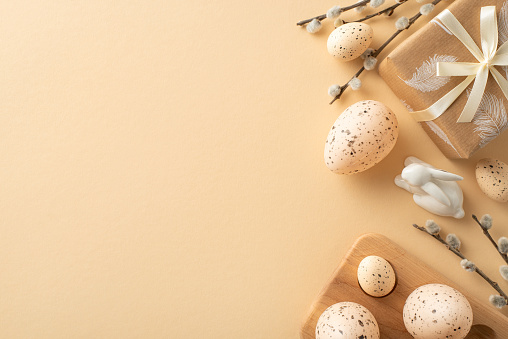 Delightful Easter tableau idea. Top view of eco-friendly wrapped present, rabbit ornament, wooden holder with quail eggs, tender pussy willow sprigs, arranged on neutral beige base with area for text