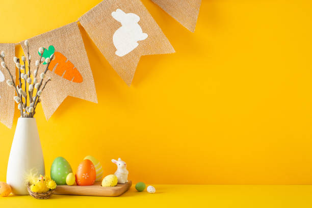 istock Easter theme setup. Picture shows side view of table decorated with rabbit, vibrant eggs, chicks, vase of pussy willow, and country-style bunting against an orange wall, leaving space for text or ads 2047995762