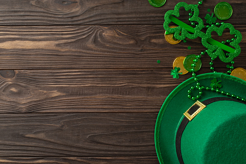 Enthralling St. Patrick's layout seen from top view, incorporating clovers, magic hat, coinage, humorous glasses, strings, confetti, neatly organized on timber background, empty spot for text or ads